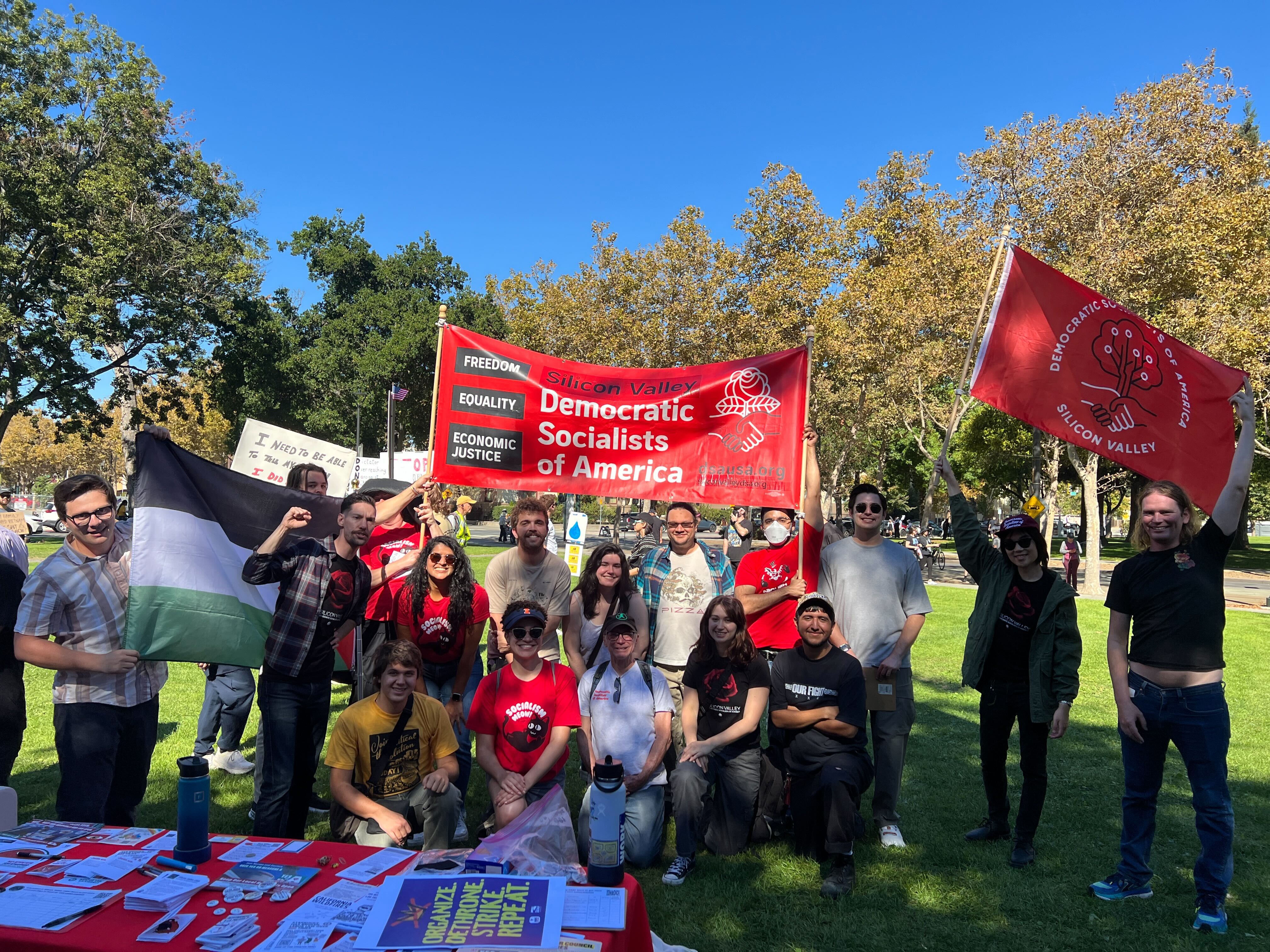 SV DSA members hold a red banner and flag while standing behind our table at the No Kings rally.