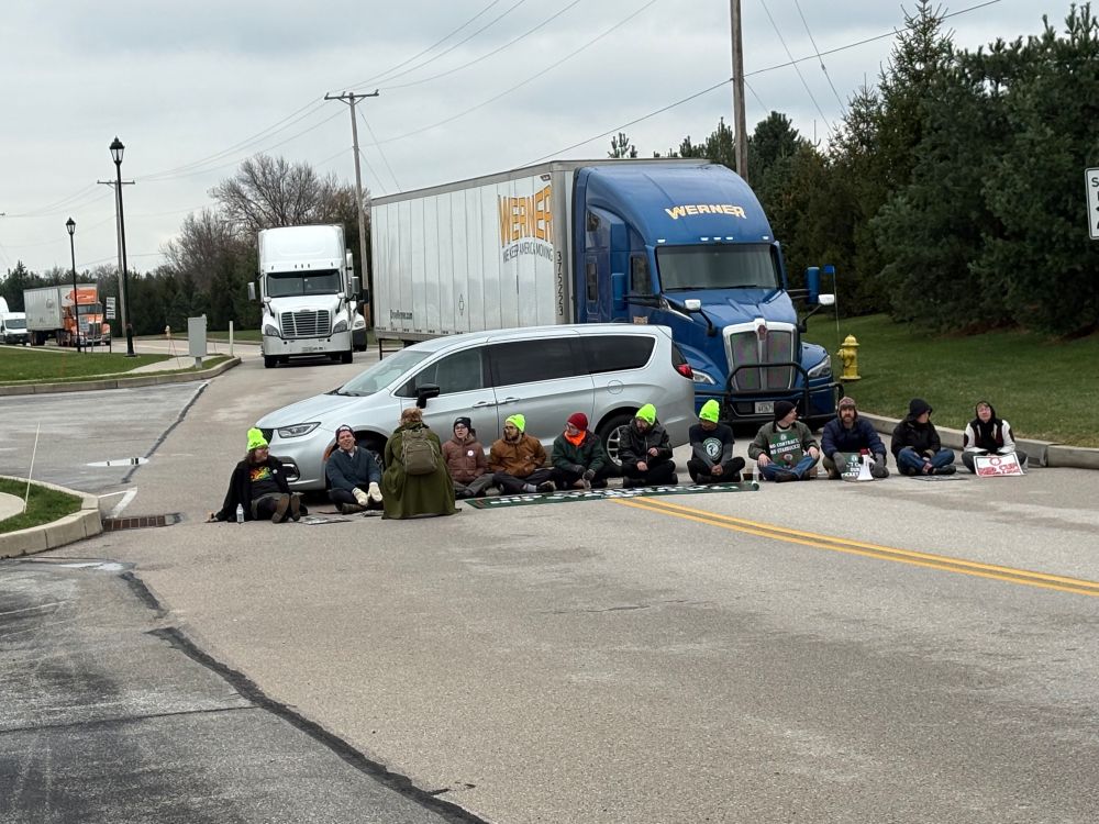 A picture of union baristas sitting down and blocking the road, huge semi trucks are stopped behind them