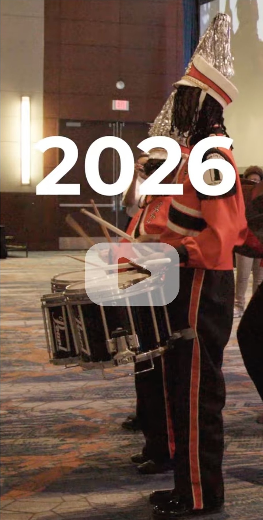 A still of a video with the word '2026' over a drummer in a marching band with a play button.