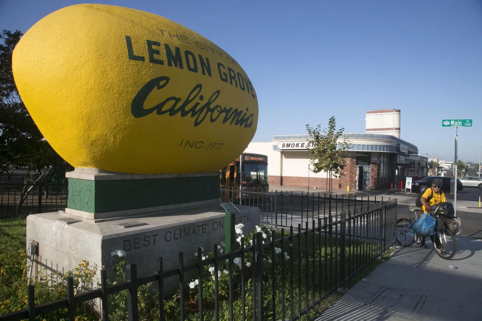 A landscape photo showing the giant lemon on Main St in Lemon Grove, with a man on a bike, the sign for Main St, and a coffee shop in the background.