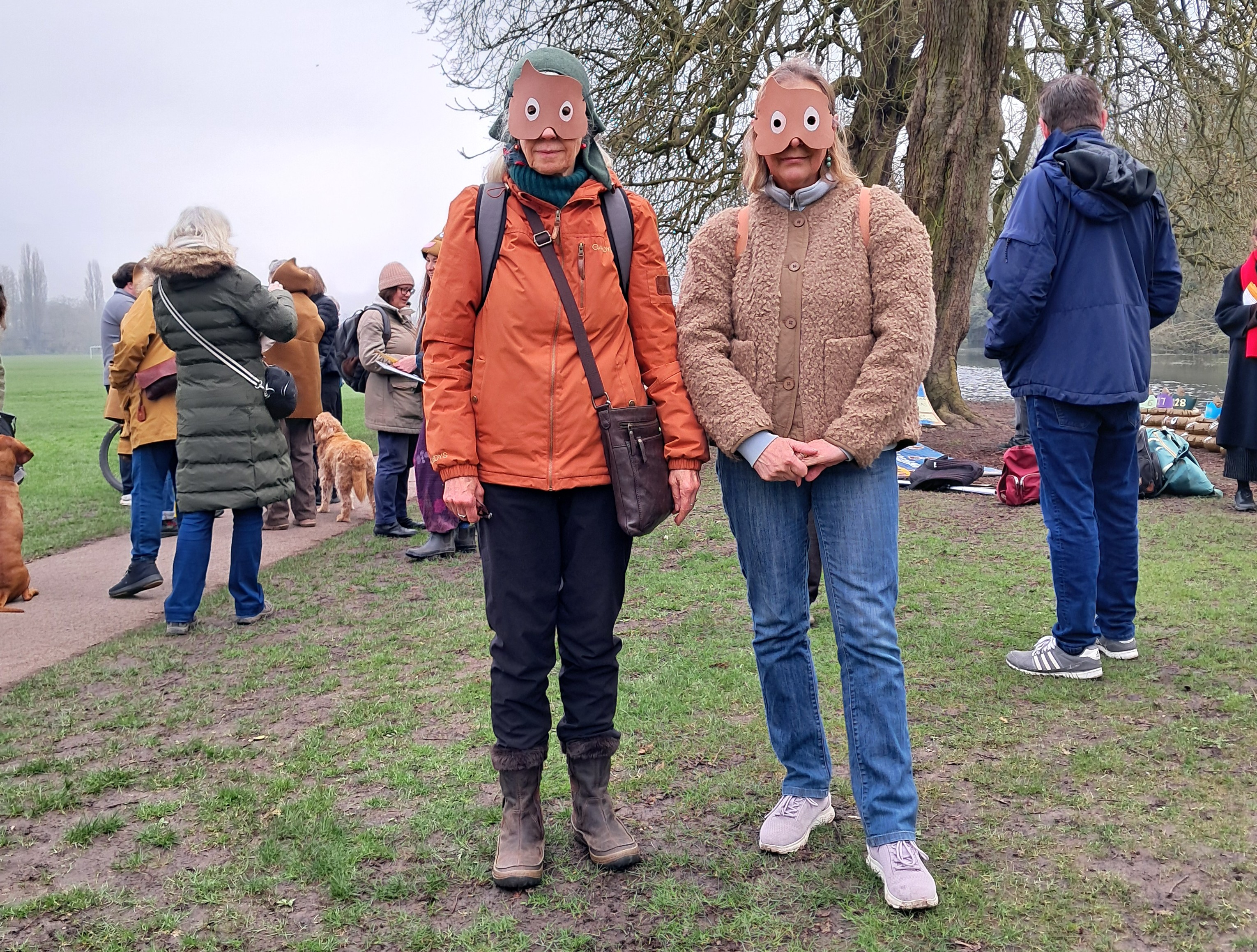 [Two people in foreground of group of people in a park wearing winter clothing.  Both are looking at the camera wearing brown masks.]