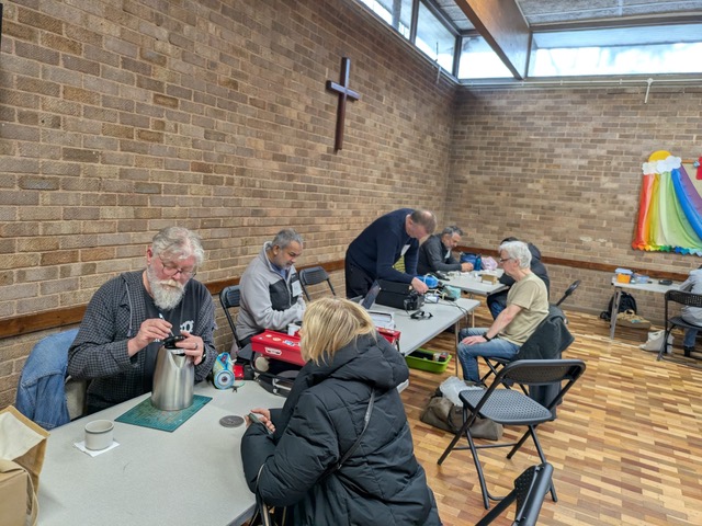 [Three tables in Christ Church Hall with repairers working on electrical equipment and owners looking on.]