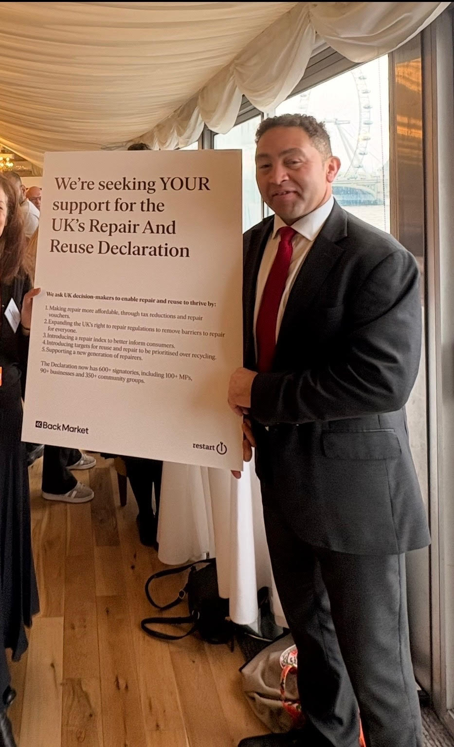 [Interior, man in dark suit and red tie (Calvin Bailey MP) holding large placard reading 
