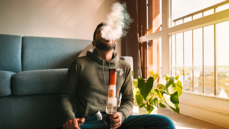 a man is seated on his living room floor next to a large window and leans against his couch smoking legal cannabis from a bong